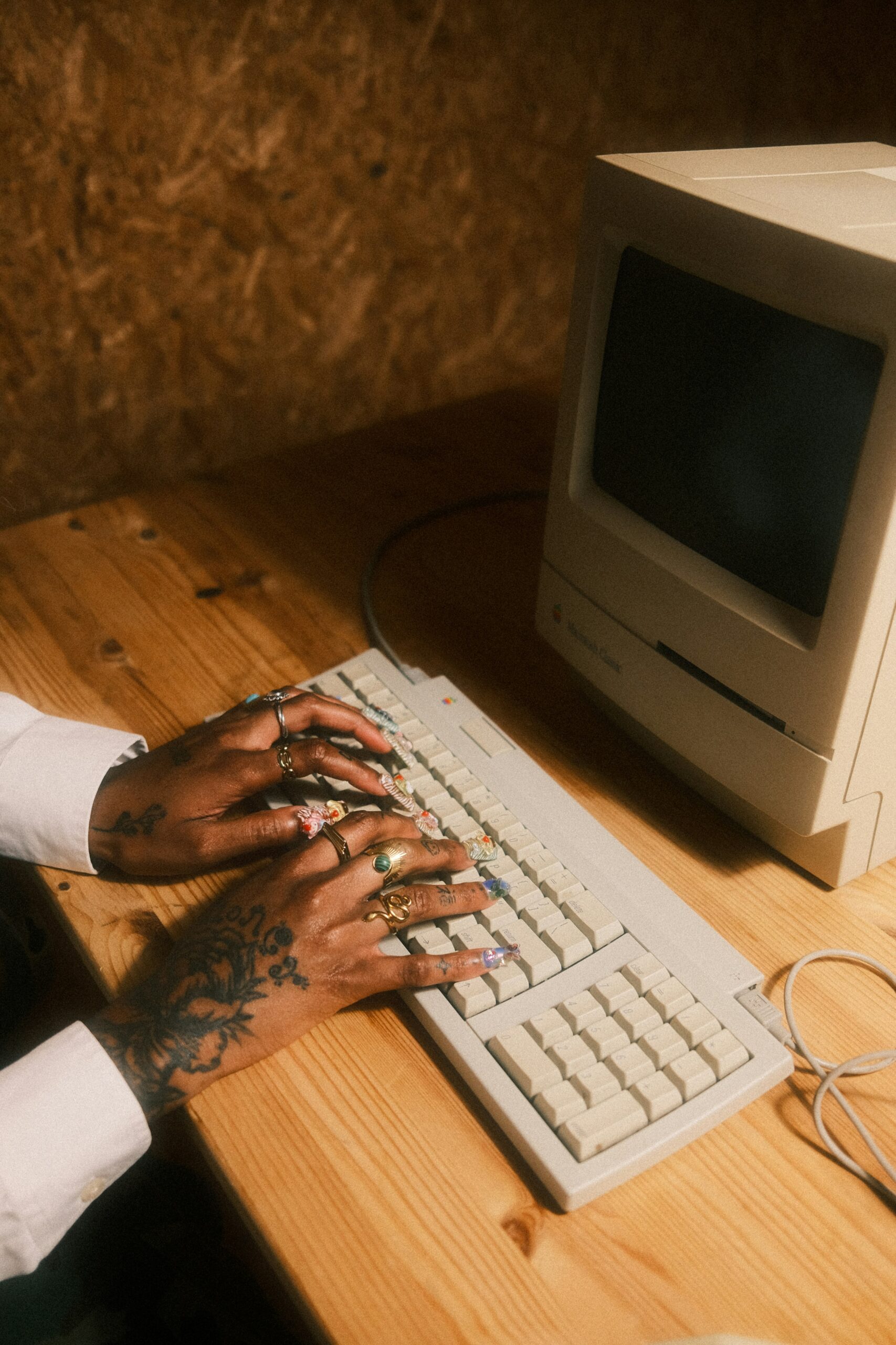 Tattooed hands type on a 1990's era desktop computer on a butcher block style wooden desktop.